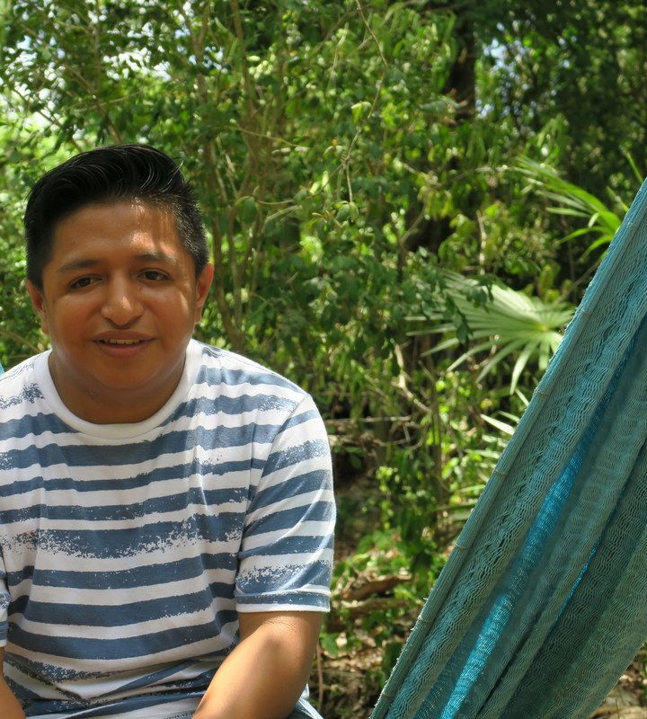 Jose sits on a blue hammock in a wooded area. He is wearing a blue and white striped shirt.