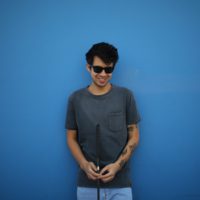 Gustavo is standing in front of a blue painted wall. He is smiling, wearing dark classes and holding his eye seeing cane.