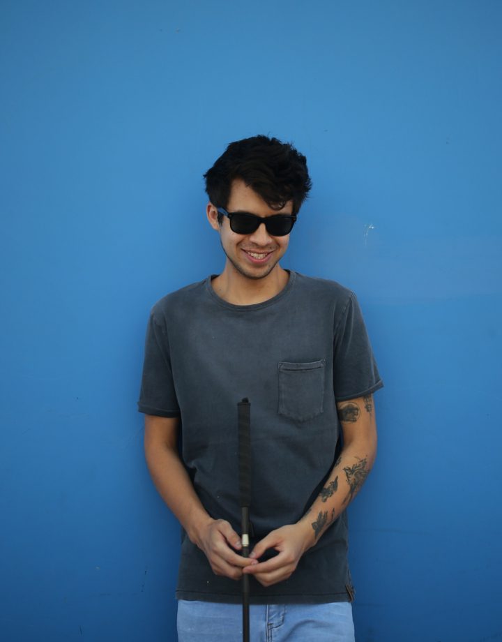 Gustavo is standing in front of a blue painted wall. He is smiling, wearing dark classes and holding his eye seeing cane.